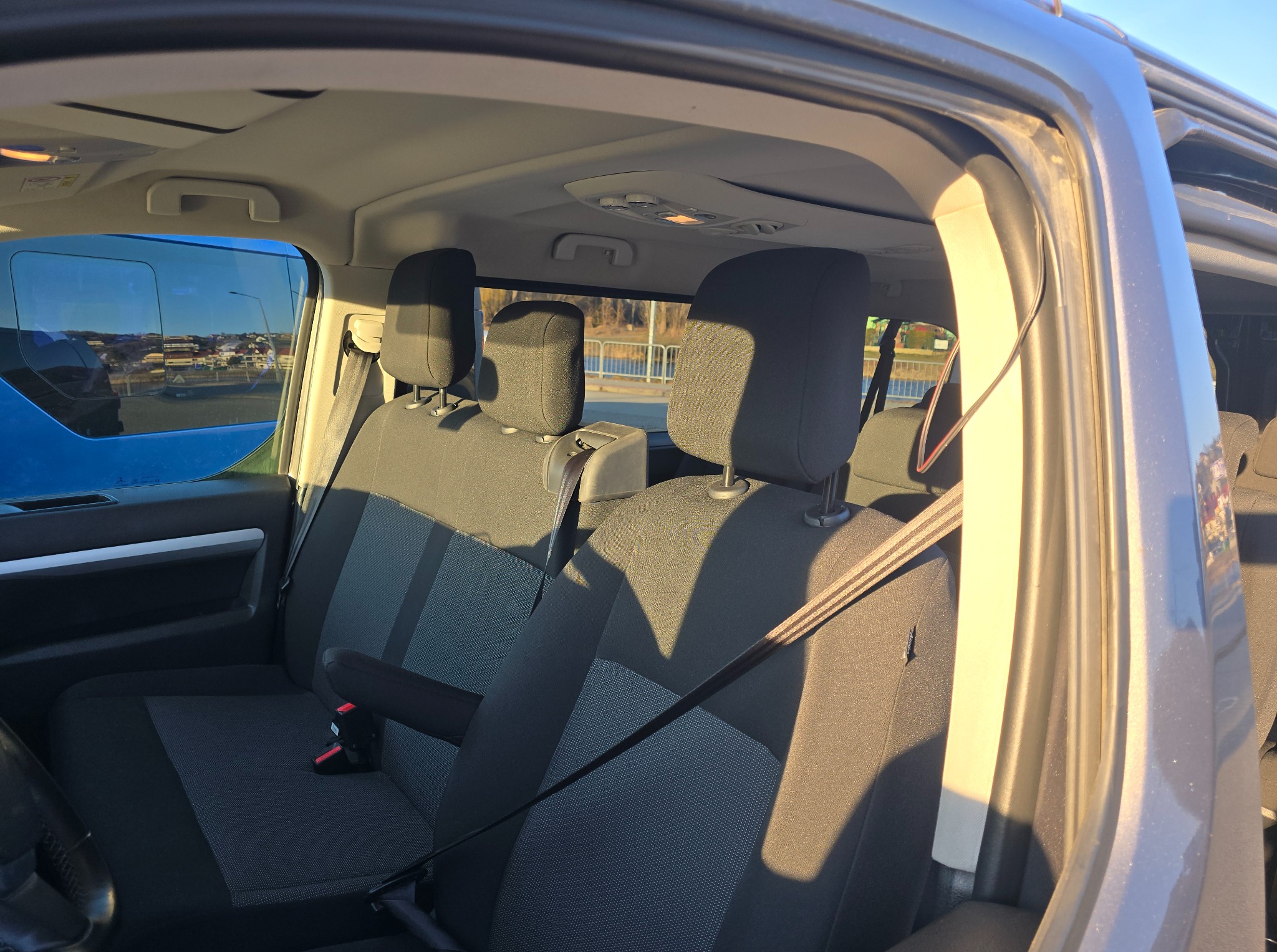 Wnętrze busa z widocznymi fotelami pasażerów, pasami bezpieczeństwa i uchwytami. Z okna widać zabudowania miejskie. Oświetlenie słoneczne podkreśla detale.