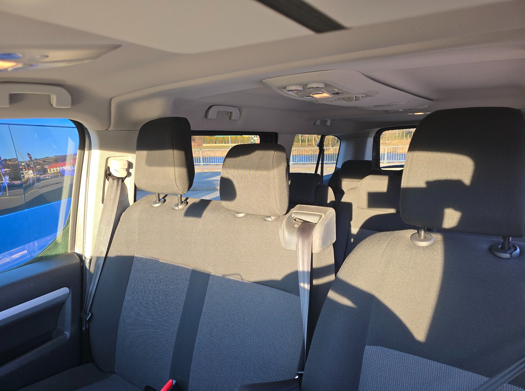 Wnętrze busa z szarymi siedzeniami i widokiem na zewnątrz przez okno. Fotel pasażera z pasem bezpieczeństwa. Oświetlenie sufitowe widoczne.