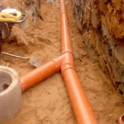 Pomarańczowe rury kanalizacyjne ułożone w wykopie ziemnym, widoczny fragment studzienki betonowej i sprzęt budowlany