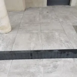 Jasnoszare płytki na schodach wejściowych w Jeleniej Górze, obramowane ciemnoszarym brukiem, prowadzące do drzwi.