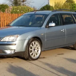 Niebieski Ford Mondeo kombi z relingami dachowymi, zaparkowany na asfalcie, z drewnianym płotem w tle.