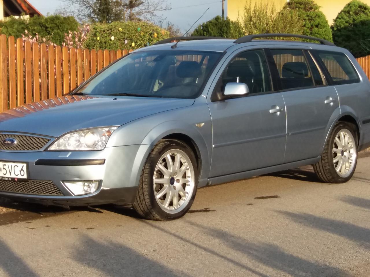 Niebieski Ford Mondeo kombi z relingami dachowymi, zaparkowany na asfalcie, z drewnianym płotem w tle.