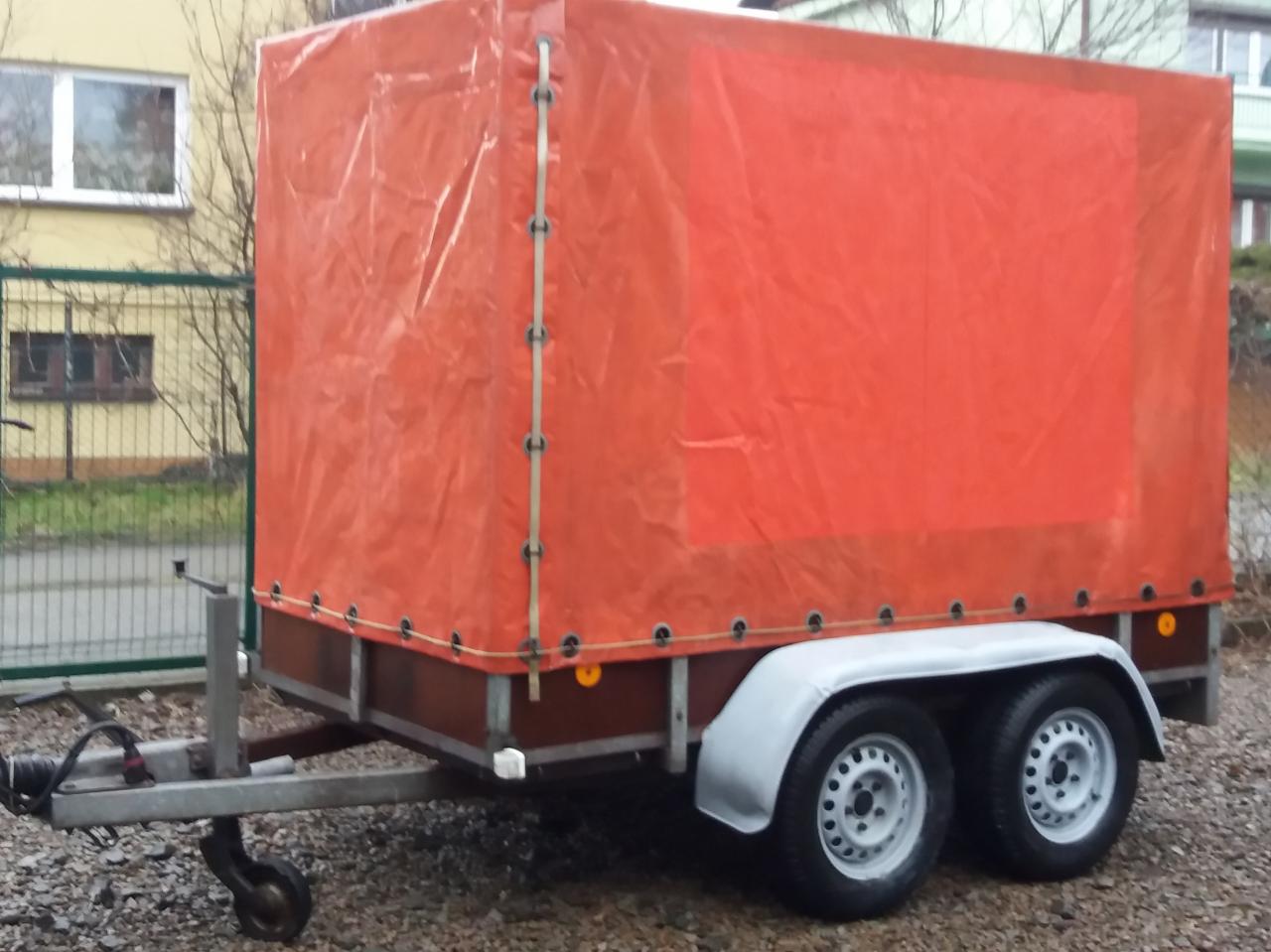 Przyczepka samochodowa z pomarańczową plandeką, widok z boku na tle budynku mieszkalnego i ogrodzenia.
