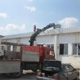 Montaż konstrukcji na dachu hali z użyciem dźwigu samochodowego, pracownik w czerwonym kombinezonie na dachu, w tle błękitne niebo z chmurami.