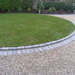 Ogród z okrągłym trawnikiem obrzeżonym szarą kostką, przechodzący w żwirową ścieżkę prowadzącą do bramy.