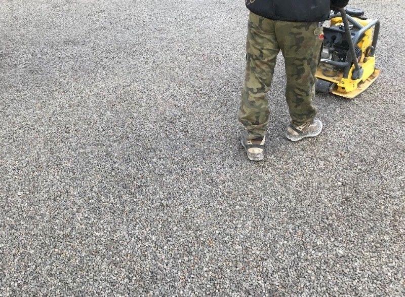 Pracownik w spodniach moro ubija zagęszczarką wibracyjną szarą nawierzchnię z drobnego kruszywa.