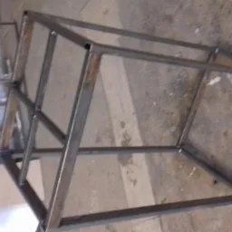 Metalowa konstrukcja klatki z kwadratowych profili, częściowo zardzewiała, widoczna w warsztacie, z elementami schodów w tle.