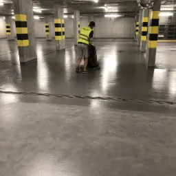Pracownik w kamizelce odblaskowej czyści posadzkę w garażu podziemnym za pomocą maszyny czyszczącej; widoczne betonowe filary z żółto-czarnymi pasami.