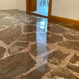 Błyszcząca, polerowana podłoga z lastryko w odcieniach brązu i bieli, z widocznym odbiciem światła z otwartego przejścia do innego pomieszczenia.