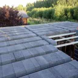 Wznoszenie stropu Teriva z szarych pustaków stropowych i belek stalowo-betonowych, widoczne zbrojenie i komin w trakcie budowy, w tle zieleń drzew.