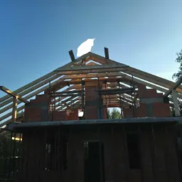 Wznoszenie konstrukcji dachu z drewnianych belek na murowanym domu w trakcie budowy, widok z dołu na tle jasnego nieba.