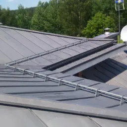 Szary dach z blachy na rąbek stojący z systemem odśnieżania w postaci rur, antena satelitarna i komin na tle zielonego krajobrazu.