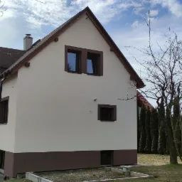 Dwukondygnacyjny dom z beżową elewacją i brązowymi detalami, widoczny z boku w słoneczny dzień, na tle błękitnego nieba i drzew bez liści.