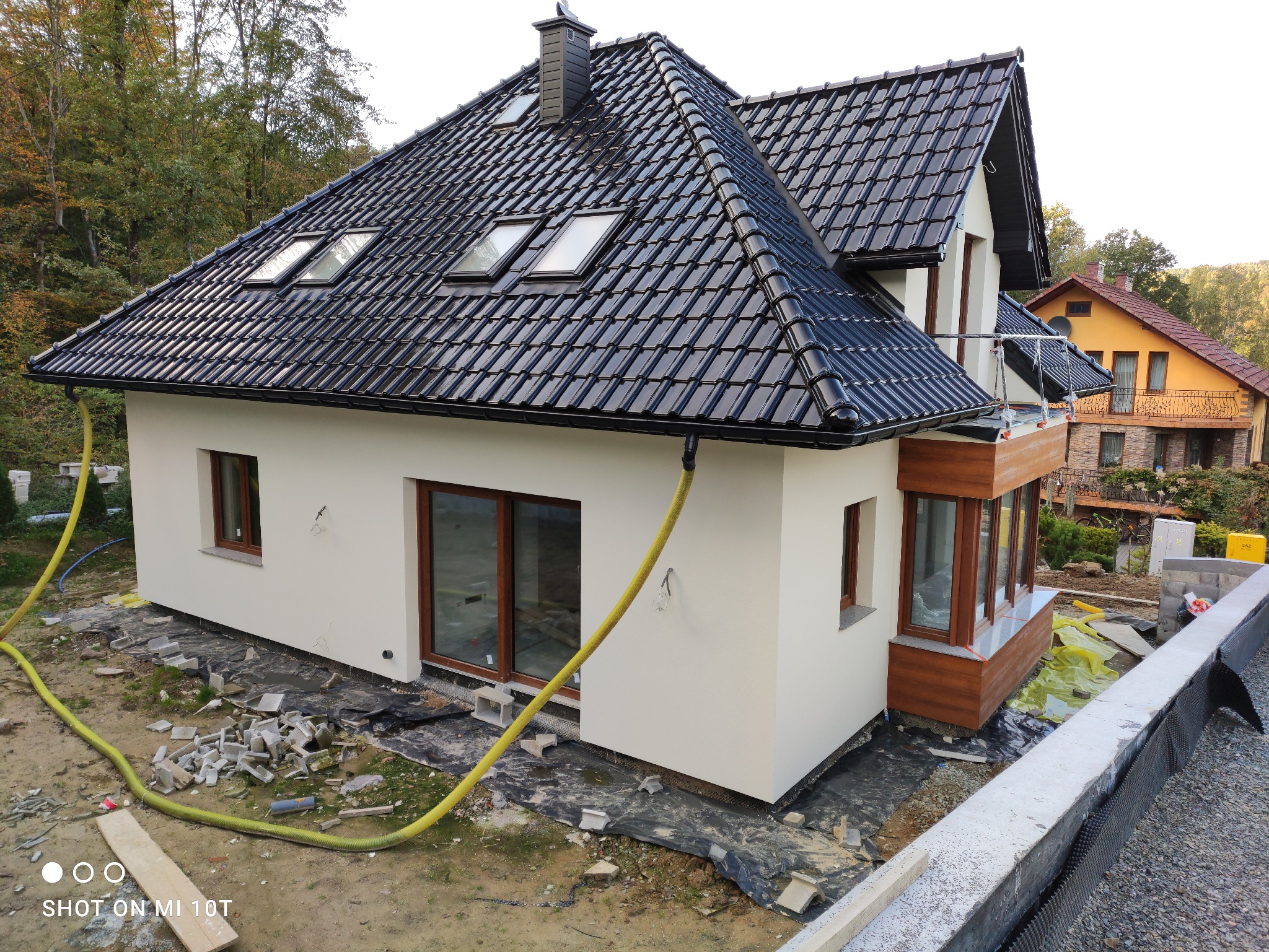 Nowoczesny dom jednorodzinny z jasną elewacją, czarną dachówką i oknami dachowymi, widoczny w trakcie prac wykończeniowych wokół budynku, z żółtym wężem ogrodowym leżącym na ziemi.