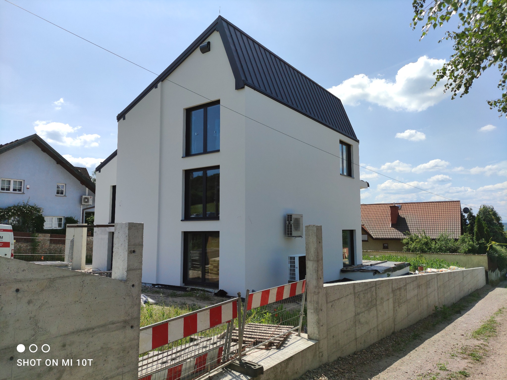 Nowoczesny, trzypiętrowy dom z białą elewacją i czarnym dachem, widoczny z perspektywy placu budowy z betonowym ogrodzeniem i barierkami ochronnymi.