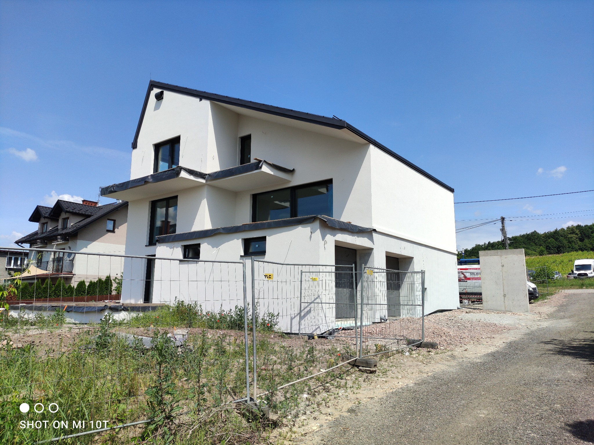 Nowoczesny dom jednorodzinny z białą elewacją i czarnymi ramami okiennymi, ogrodzony tymczasowym płotem budowlanym na tle błękitnego nieba.