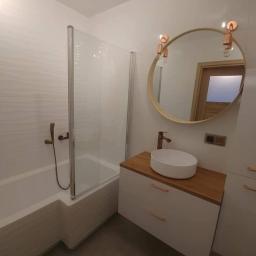 Marek Foltyn Usługi Wykończeniowe - Nowoczesna łazienka z wanną za szklaną ścianką, okrągłym lustrem z miedzianymi lampami i białą szafką z drewnianym blatem oraz miedzianą armaturą.