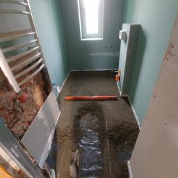 Marek Foltyn Usługi Wykończeniowe - Łazienka w trakcie remontu: ściany wyłożone płytami karton-gips, wylewka na podłodze z folią i poziomnicą, grzejnik drabinkowy w narożniku, widoczne narzędzia murarskie.