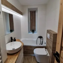 Nowoczesna łazienka z białymi i drewnopodobnymi płytkami, owalną umywalką na drewnianym blacie, wiszącą toaletą i wanną z obudową z płytek drewnopodobnych.