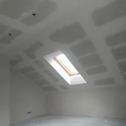 Poddasze ze skośnym sufitem, na którym widać świeżo położoną płytę kartonowo-gipsową, przygotowaną do dalszych prac wykończeniowych, okno dachowe wpuszcza naturalne światło.