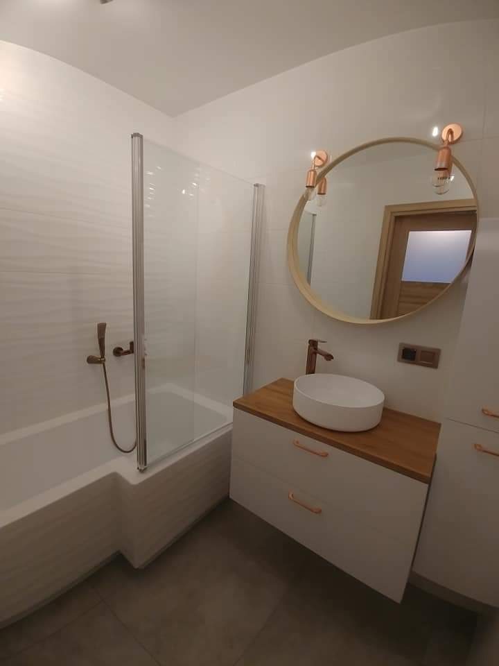 Nowoczesna łazienka z wanną za szklaną ścianką, okrągłym lustrem z miedzianymi lampami i białą szafką z drewnianym blatem oraz miedzianą armaturą.