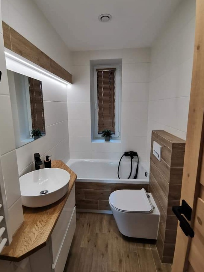 Nowoczesna łazienka z białymi i drewnopodobnymi płytkami, owalną umywalką na drewnianym blacie, wiszącą toaletą i wanną z obudową z płytek drewnopodobnych.