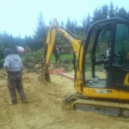 Żółta mini koparka JCB 8018 CTS na placu budowy z widocznymi pracownikami, wykopem i ułożoną rurą kanalizacyjną w tle domu jednorodzinnego.