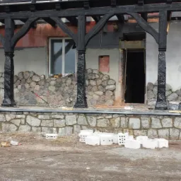 Montaż kamiennej elewacji na tarasie z czarną drewnianą konstrukcją zadaszenia, widoczne białe bloczki i narzędzia.