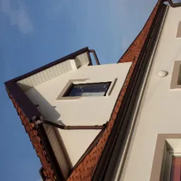 Fragment elewacji budynku z oknami o geometrycznych obramowaniach, brązowa dachówka i rynny na tle błękitnego nieba.