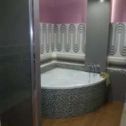 Wykończona łazienka z narożną wanną obudowaną mozaiką, kabiną prysznicową z szarymi drzwiami i ścianami w odcieniach różu i szarości z dekoracyjnymi płytkami.