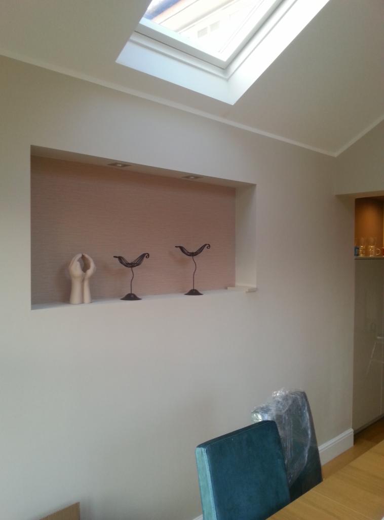 Pomalowane na jasny kolor ściany z wnęką, w której znajdują się dekoracje: rzeźba dłoni i dwie metalowe figurki ptaków. Na suficie widoczne okno dachowe.