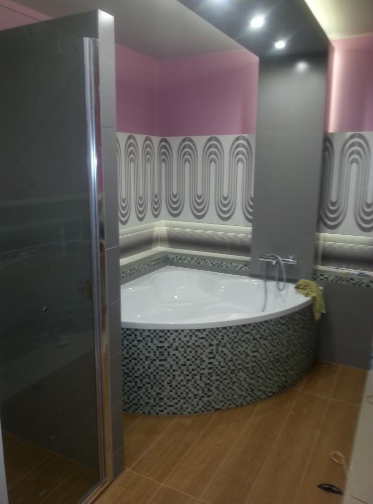 Wykończona łazienka z narożną wanną obudowaną mozaiką, kabiną prysznicową z szarymi drzwiami i ścianami w odcieniach różu i szarości z dekoracyjnymi płytkami.