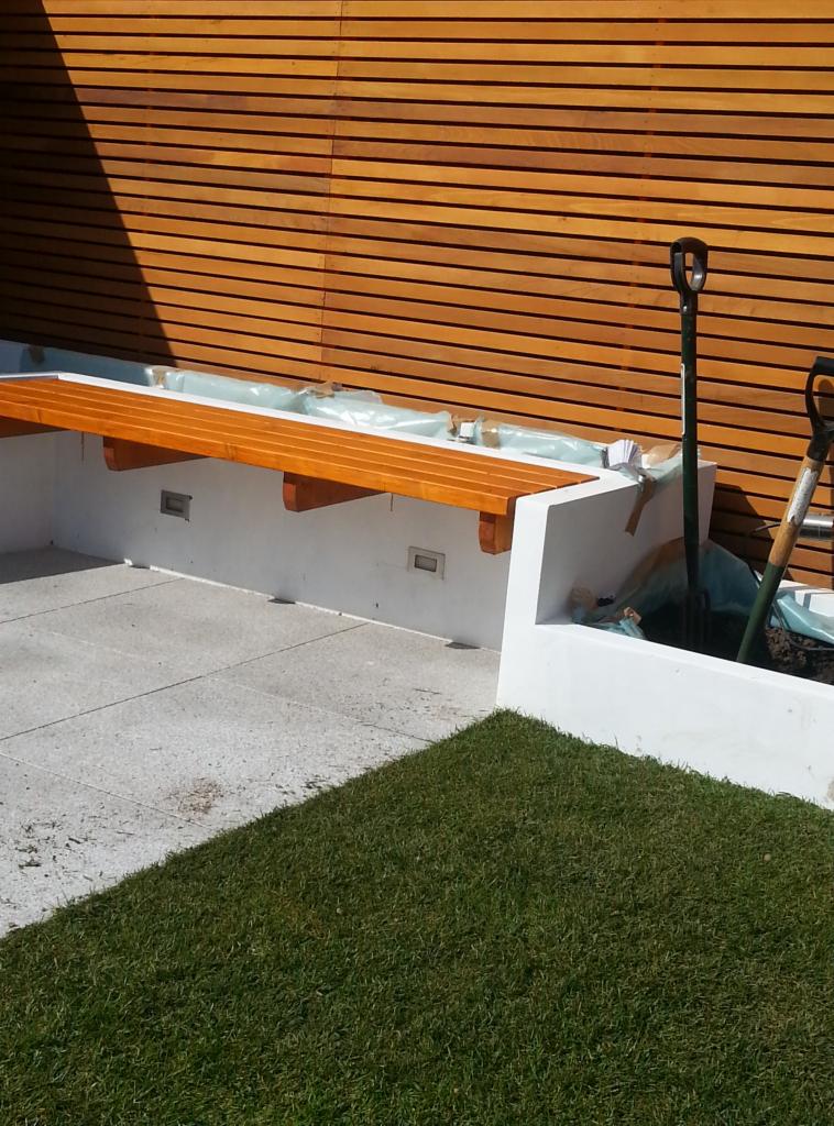 Nowoczesny ogród z drewnianą ścianą, betonową ławką z podświetleniem i świeżo posianą trawą, widoczne narzędzia ogrodnicze.