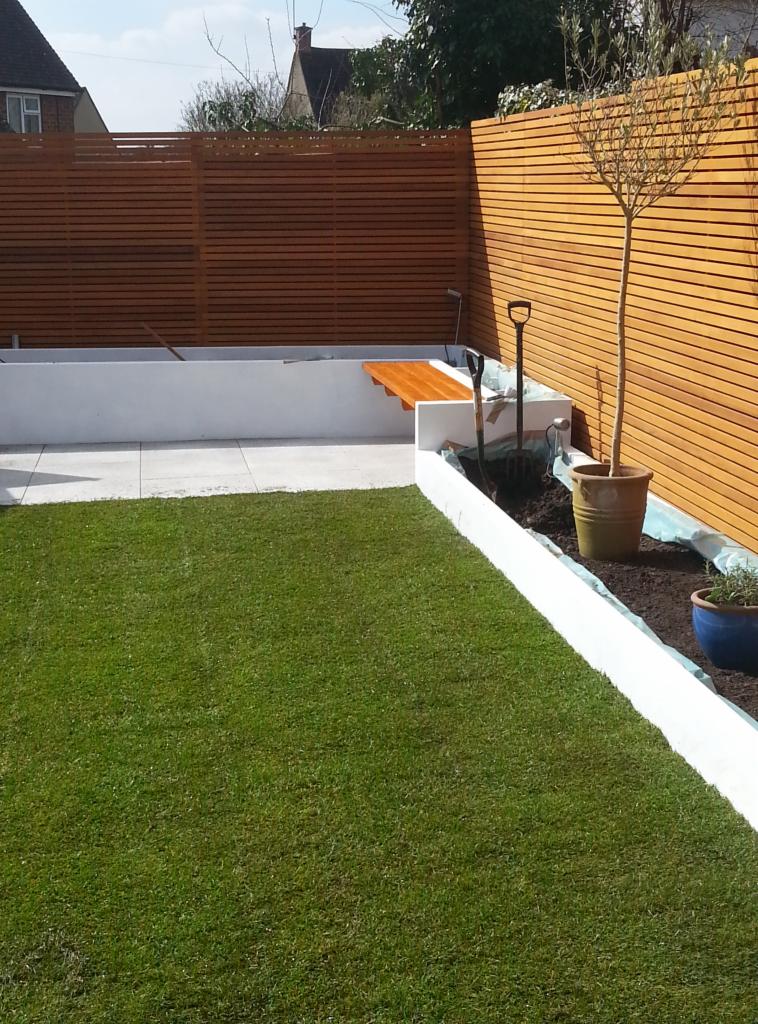 Nowoczesny ogród z drewnianym ogrodzeniem panelowym, świeżo posiana trawa, białe obrzeża rabat, narzędzia ogrodnicze i doniczki w trakcie aranżacji.