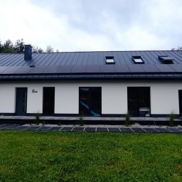 RESS-INWESTYCJE SPÓŁKA Z OGRANICZONĄ ODPOWIEDZIALNOŚCIĄ - Biały dom z ciemnym dachem i panelami słonecznymi, widok z trawnika. Nowoczesna architektura z elementami OZE.