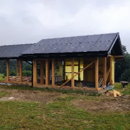 Montaż paneli słonecznych na drewnianej konstrukcji budynku w trakcie budowy. Widoczne panele, drewniane ramy i elementy instalacji. Tło: zielona łąka i las.