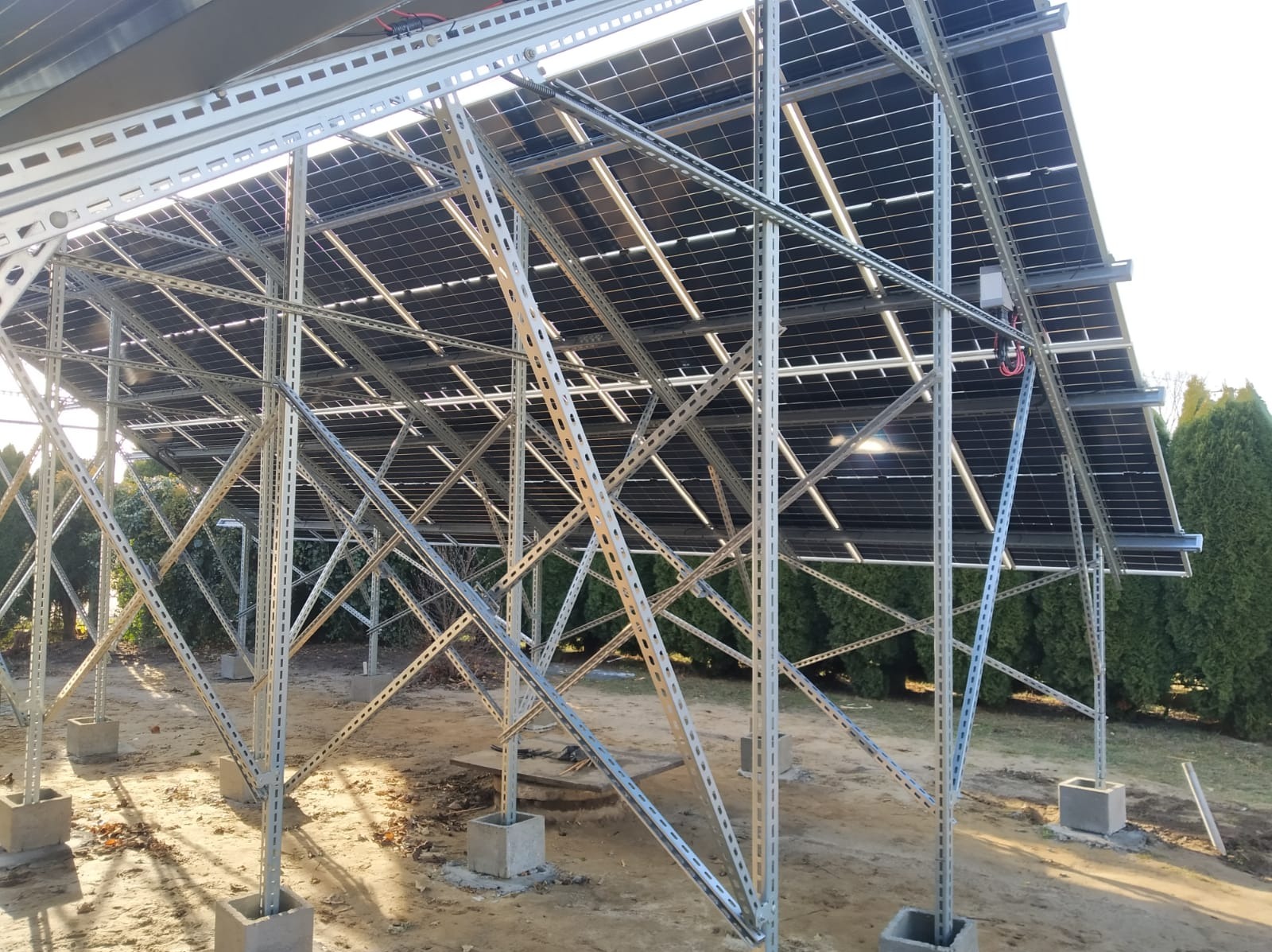Instalacja fotowoltaiczna na gruncie w Przeworsku, widok od spodu na metalową konstrukcję wsporczą z betonowymi podstawami, panele ustawione pod kątem, widoczne krzewy w tle.
