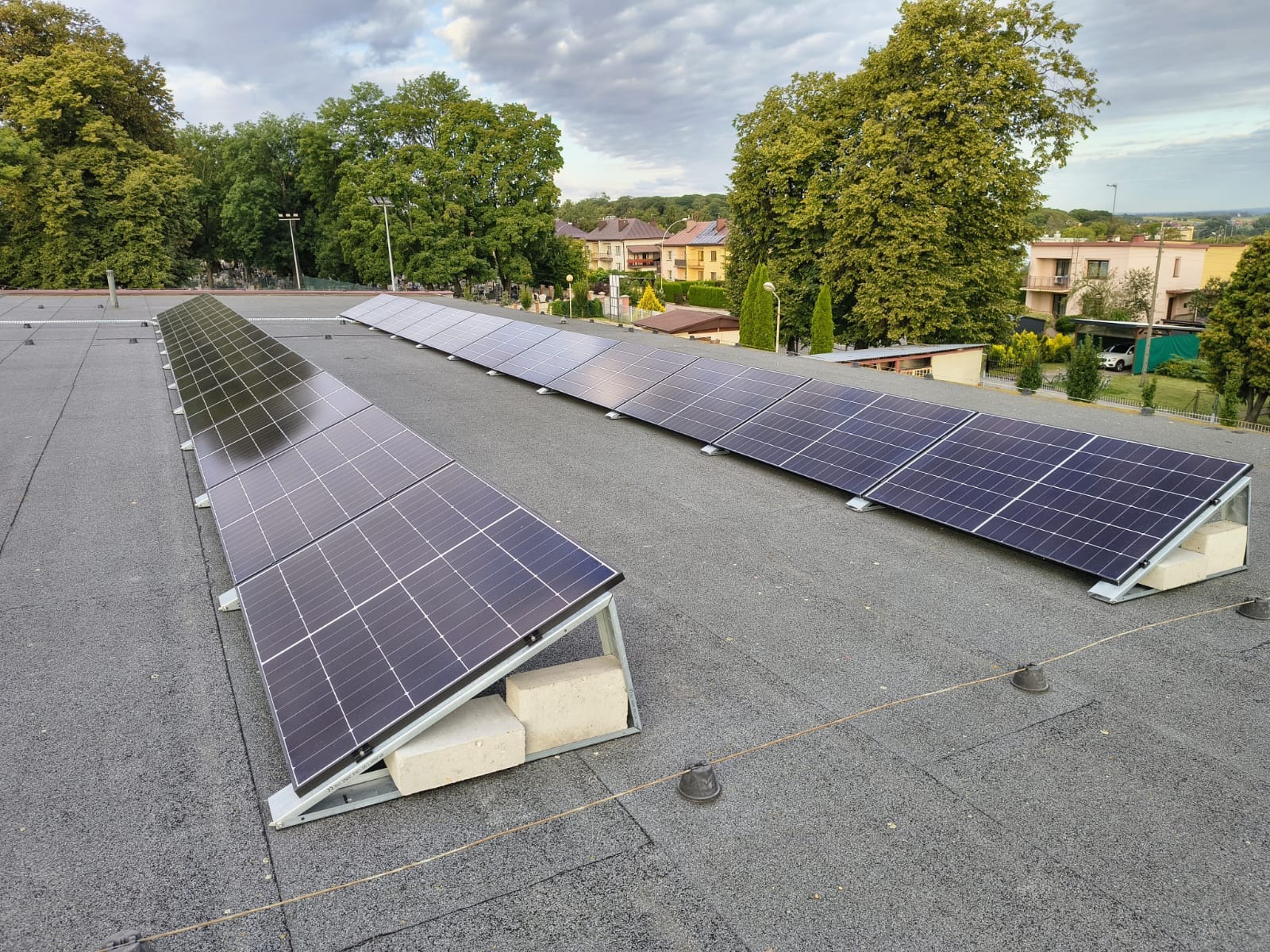 Instalacja paneli słonecznych na płaskim dachu budynku, z widocznymi blokami betonowymi jako obciążenie, w tle zabudowa mieszkalna i zieleń.