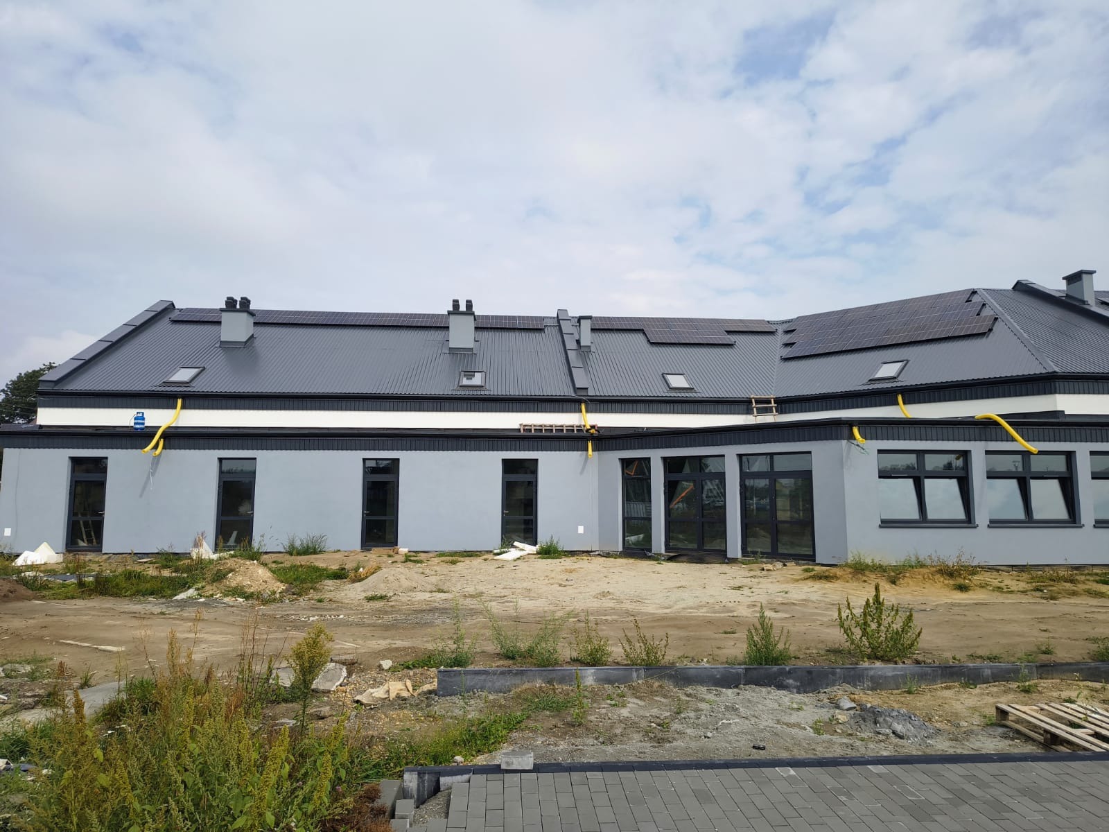 Szary budynek z panelami słonecznymi na dachu, widoczne żółte rury i drabina. Teren wokół budynku w trakcie prac wykończeniowych.
