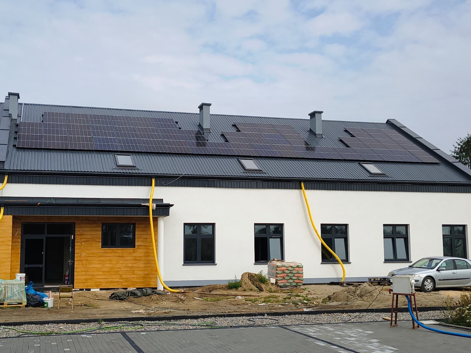 Dom z panelami słonecznymi na dachu i żółtymi rurami spustowymi. Widoczne okna dachowe, cegły i samochód. Ujęcie z zewnątrz budynku.