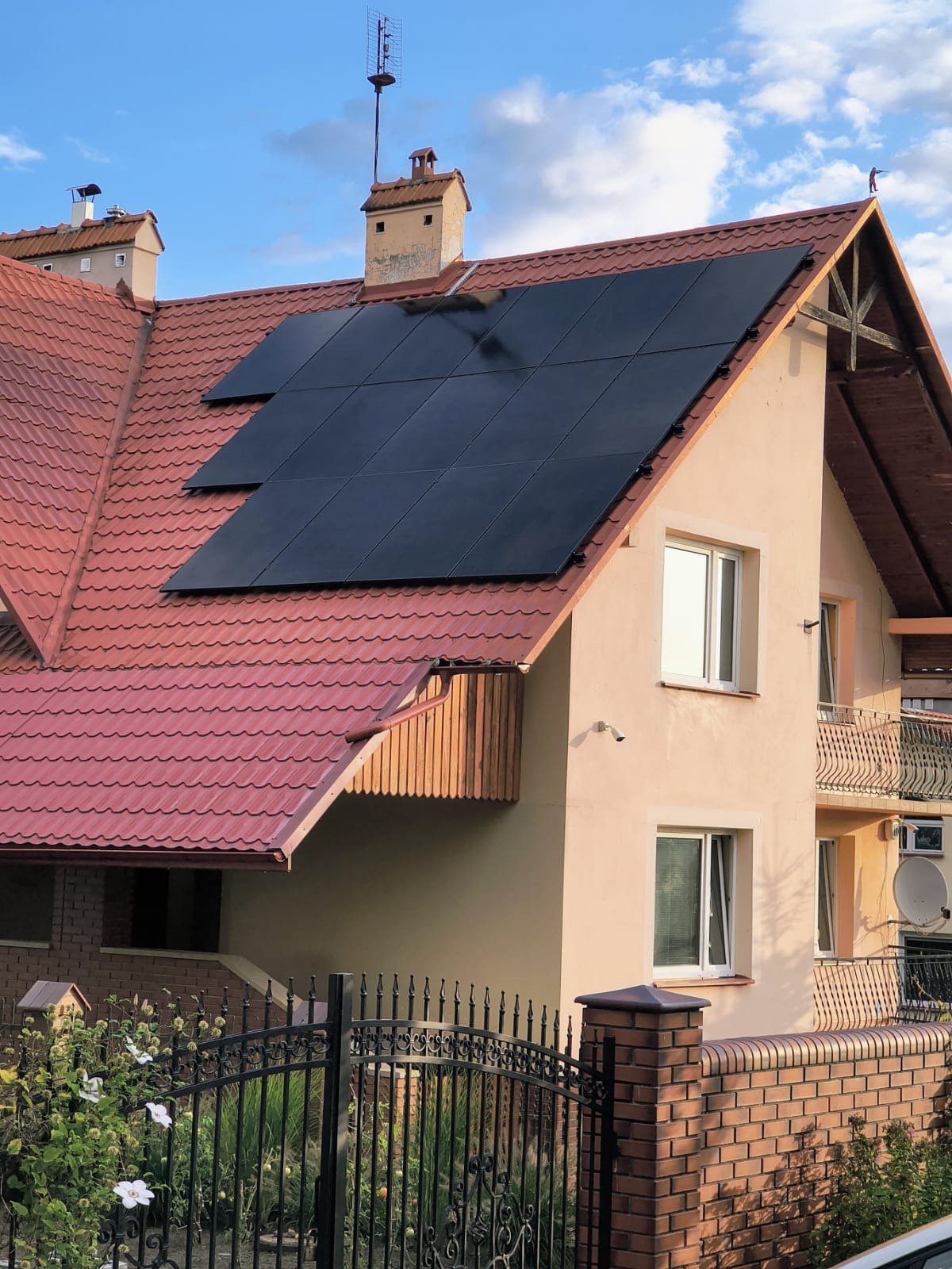 Ciemne panele na czerwonym dachu domu jednorodzinnego z anteną. Widok z dołu, błękitne niebo z chmurami w tle. Ogród z kutym ogrodzeniem na pierwszym planie.