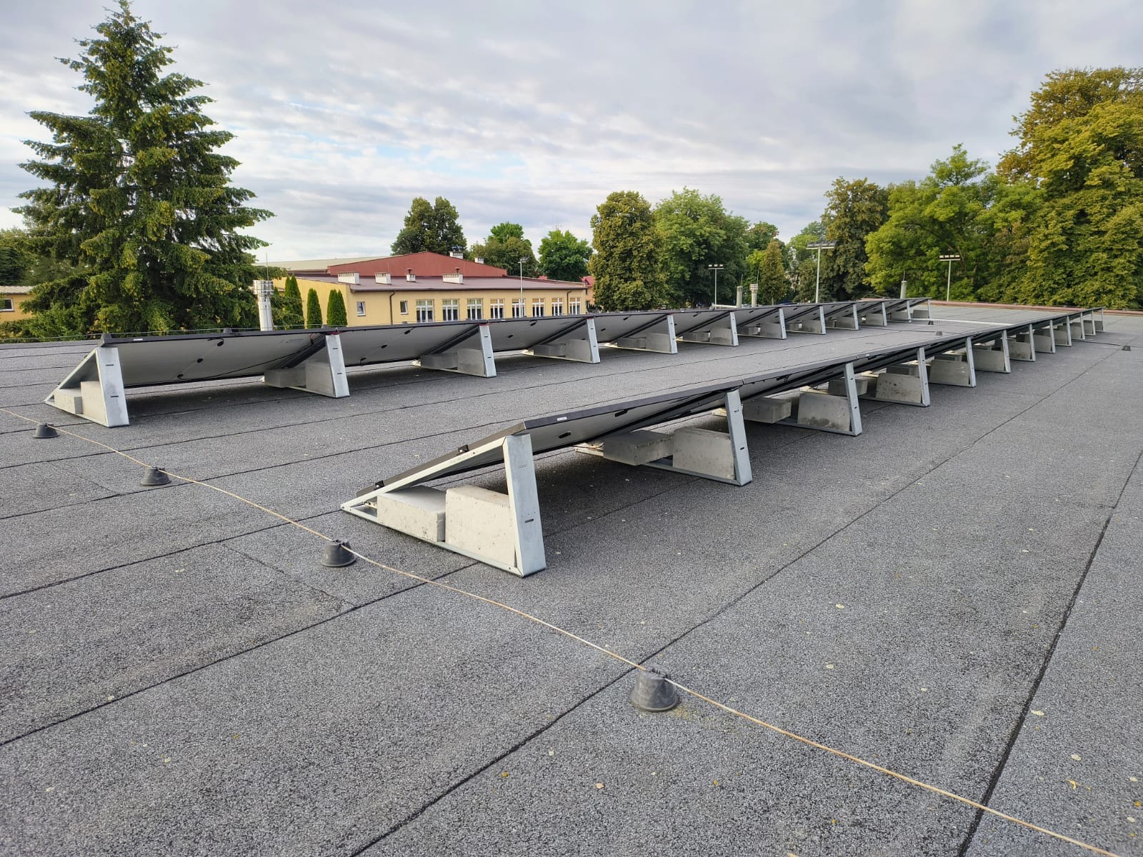 Rząd paneli słonecznych na płaskim dachu z betonowymi podstawami, widok z perspektywy, budynek w tle, pochmurne niebo.