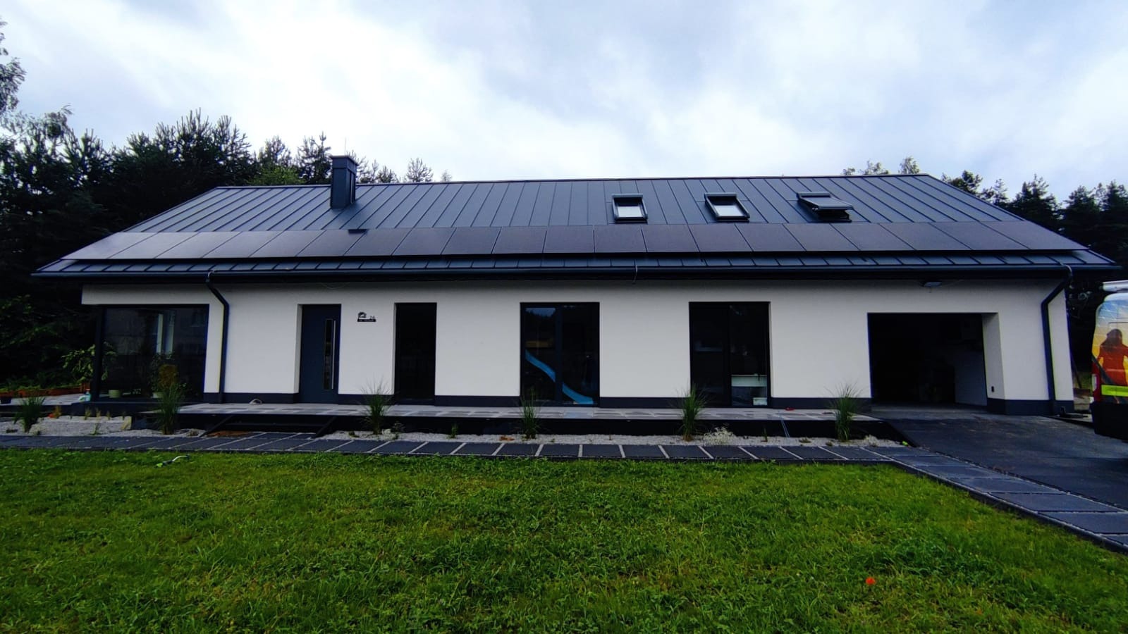 Biały dom z ciemnym dachem i panelami słonecznymi, widok z trawnika. Nowoczesna architektura z elementami OZE.