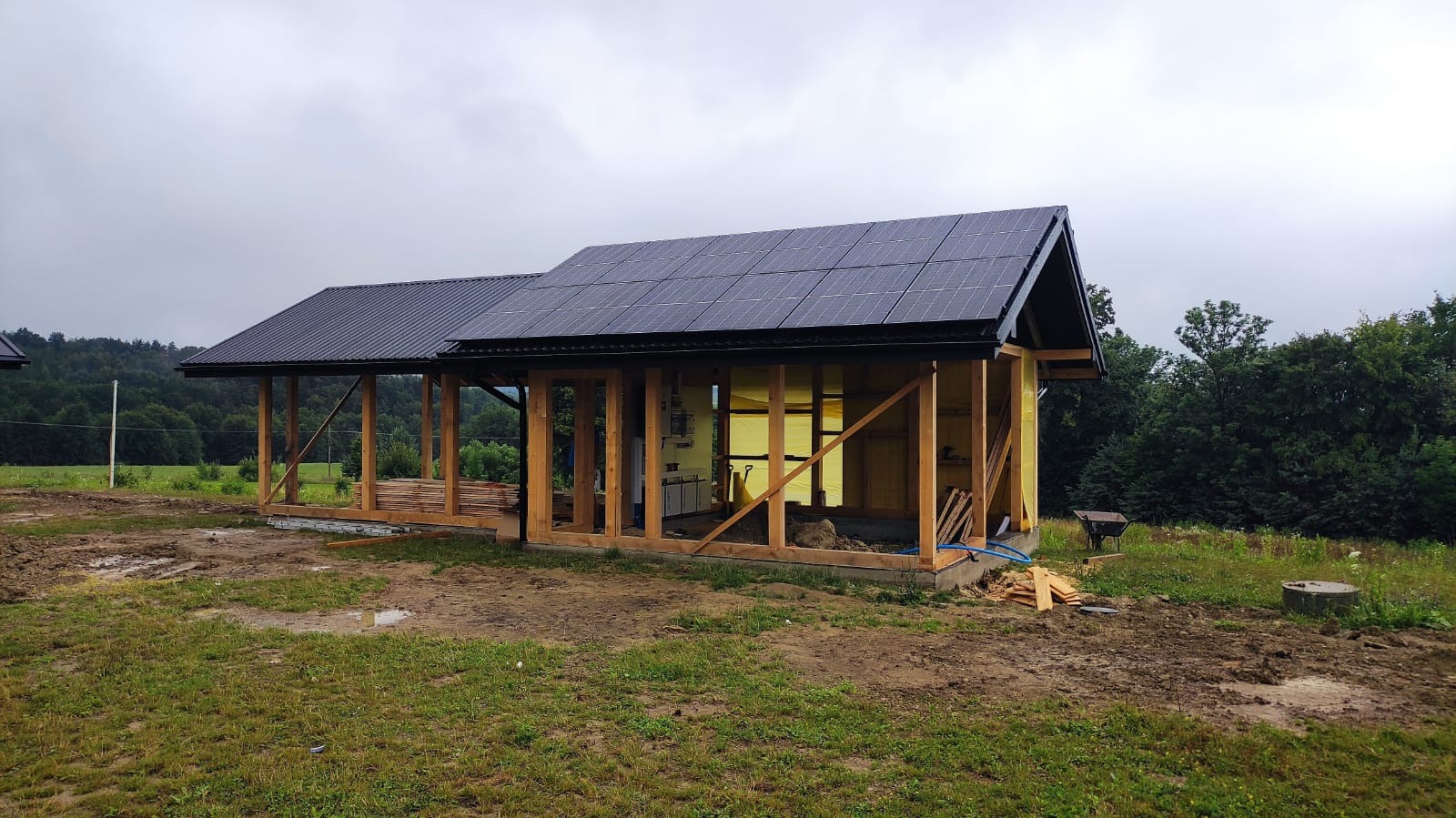 Montaż paneli słonecznych na drewnianej konstrukcji budynku w trakcie budowy. Widoczne panele, drewniane ramy i elementy instalacji. Tło: zielona łąka i las.