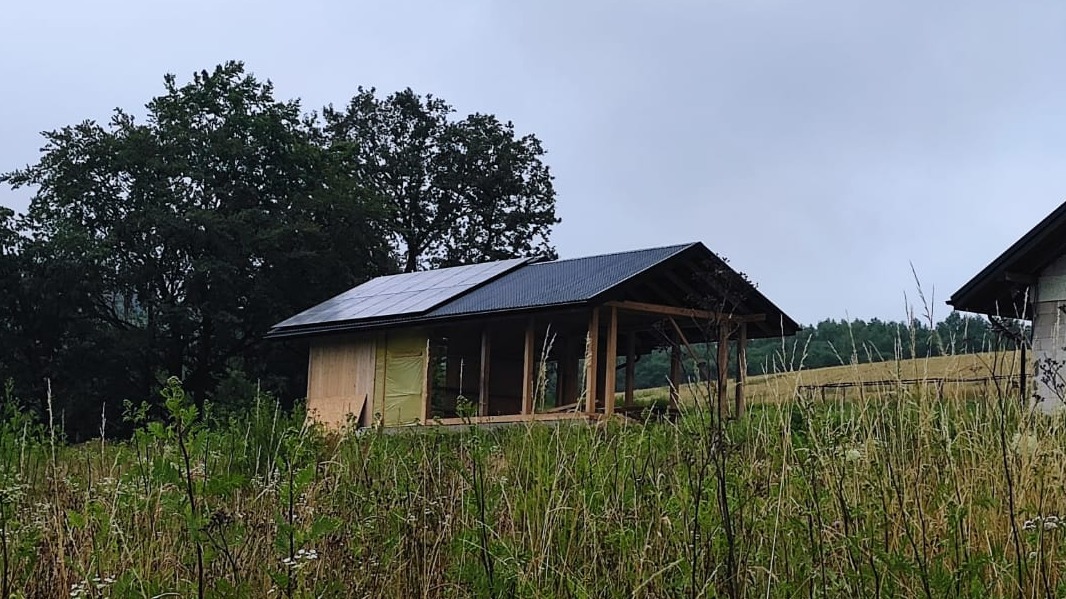 Niedokończona konstrukcja z panelami słonecznymi na dachu, otoczona wysoką trawą i drzewami, w pochmurny dzień. Widoczny szkielet budynku i częściowo zamontowane ściany.