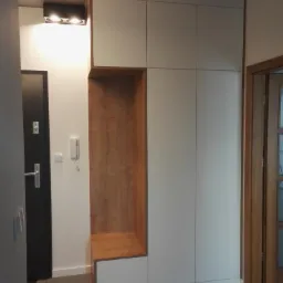 Wysoka szafa w przedpokoju z białymi frontami i drewnianą wnęką z siedziskiem, minimalistyczny design, widoczny betonowy sufit i czarne drzwi wejściowe.