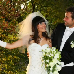 Para młoda w plenerze: Panna młoda w sukni bez ramiączek, z welonem i bukietem białych róż, opiera dłoń o korę drzewa, spoglądając z uśmiechem na pana młodego w czarnym smokingu z białą muchą.