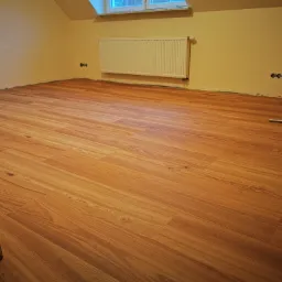 Jasnobrązowe panele podłogowe ułożone w pokoju na poddaszu, widoczny grzejnik i listwa przypodłogowa wzdłuż ściany.