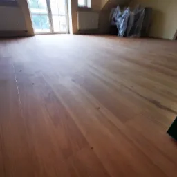 Nowo ułożona, lakierowana podłoga drewniana w jasnym, przestronnym pokoju na poddaszu, z oknami i grzejnikami w tle, meble zabezpieczone folią malarską.