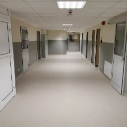 Korytarz w obiekcie publicznym z jasną podłogą z wykładziny PCV, białe drzwi z czarnymi obramowaniami, ściany dwukolorowe szaro-beżowe, oświetlenie sufitowe.